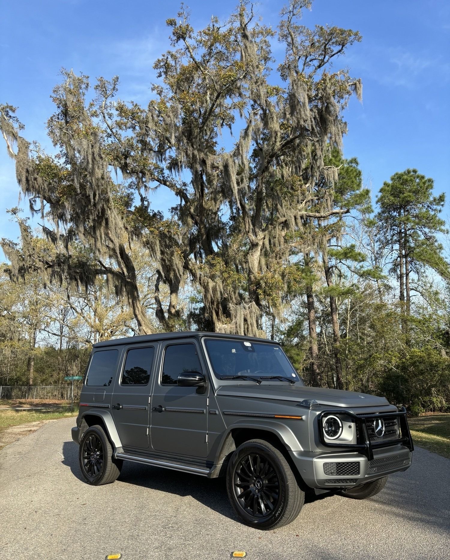 Gray Mercedes G-Wagon SUV parked on a road under a large tree with Spanish moss. Sunny day.