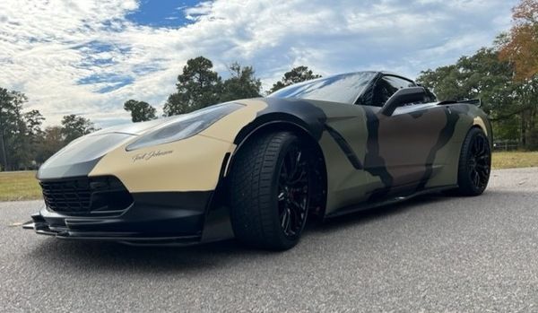 Camouflage-wrapped black Corvette sports car on a paved road with a cloudy sky and trees.