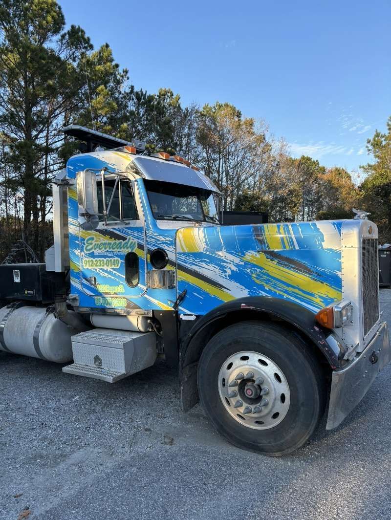 Blue and yellow semi-truck with faded paint, parked outdoors on gravel, under a blue sky.