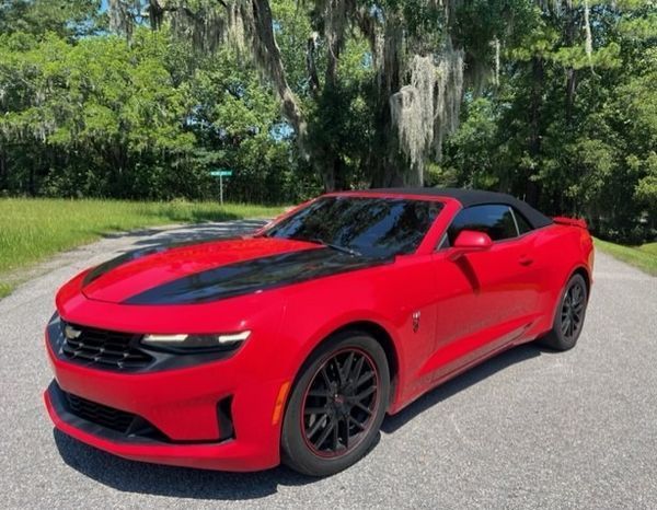 Red Chevrolet Camaro convertible with black racing stripes on a tree-lined road.