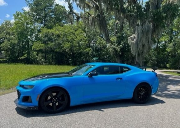Bright blue Chevrolet Camaro sports car parked on a road with a black hood, wheels, and spoiler, in a green outdoor setting.