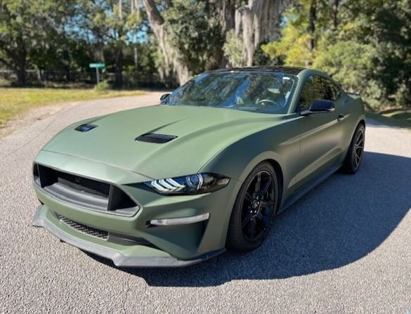Green matte-finish Ford Mustang on a road, parked near trees on a sunny day.