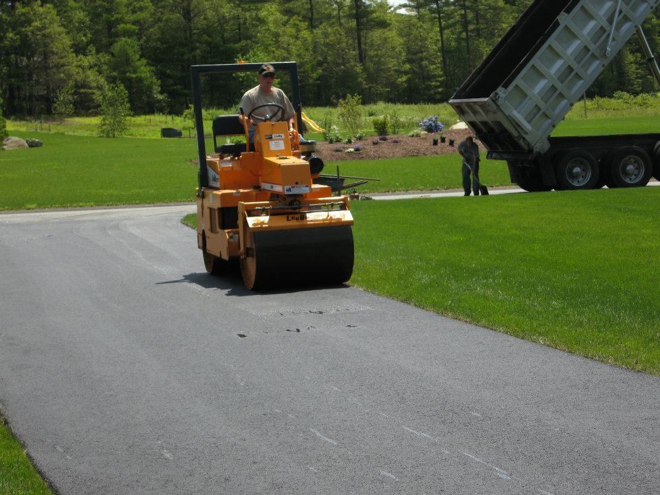 A man is driving a roller down a road next to a dump truck