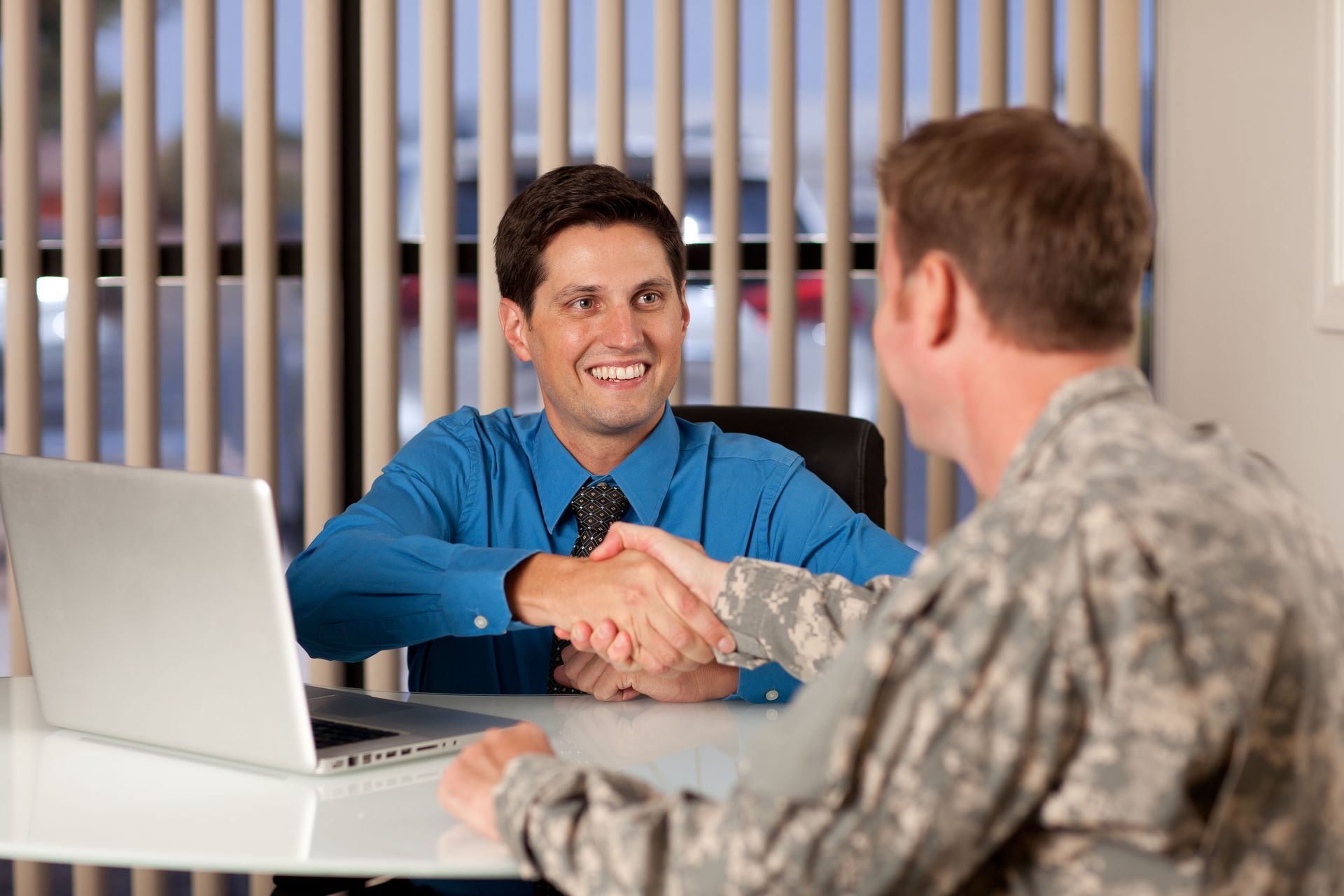 Two people shaking hands across a desk in a bright office, with a laptop nearby.