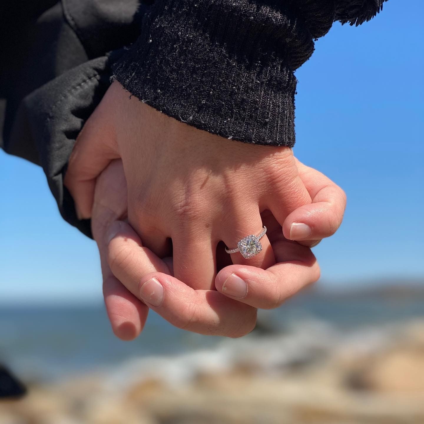 Two hands holding, one with a diamond engagement ring, against a blue sky and ocean background.