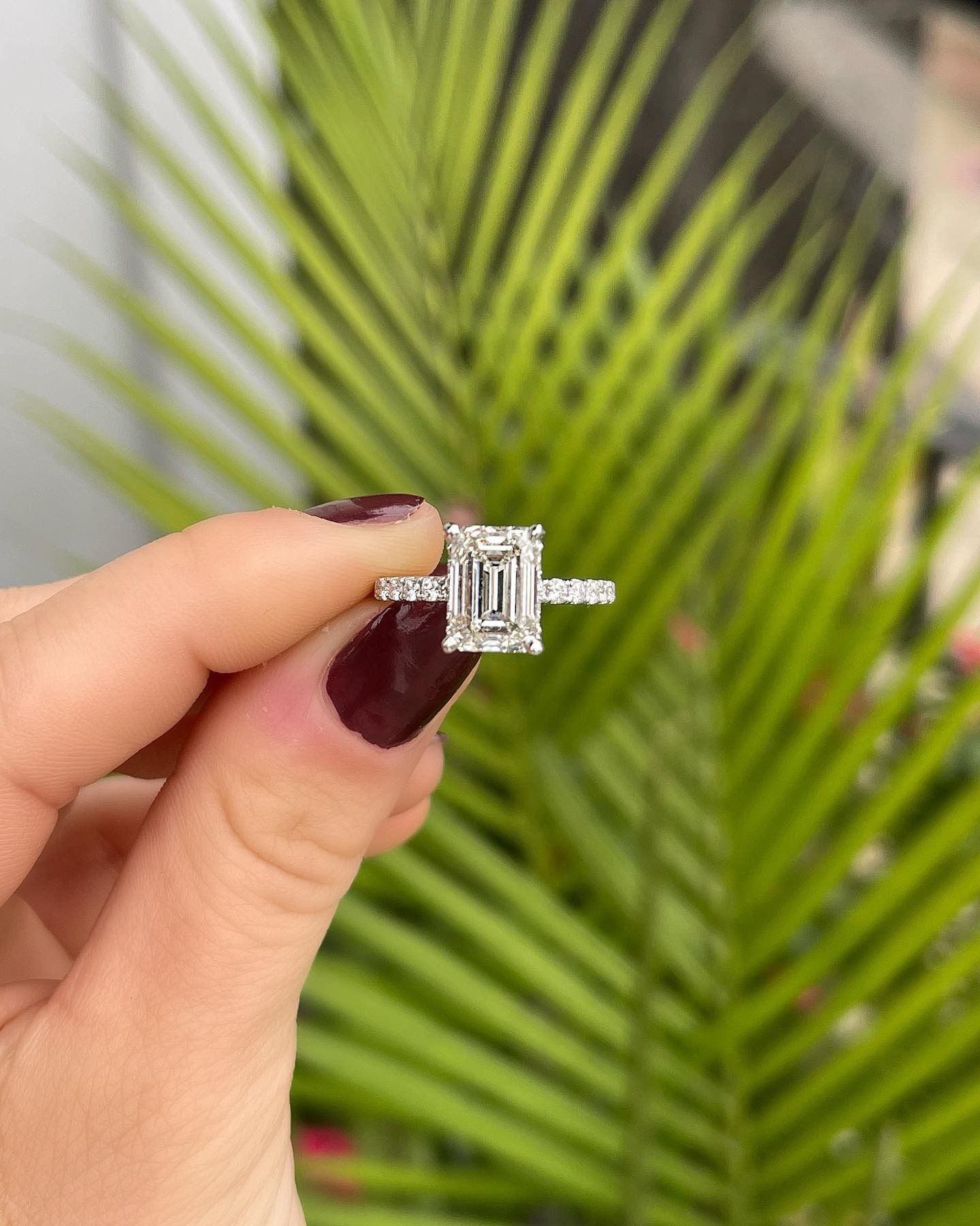 Woman's hand holding a diamond engagement ring with emerald cut center stone. Green leaves in the background.
