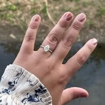 Hand with an oval-cut diamond ring.  The person has light skin, wearing a floral-print sleeve, near water.