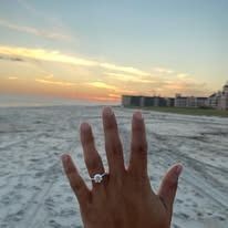 Hand with engagement ring held up against a beach sunset.