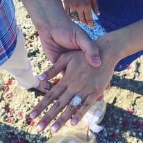 Hands holding, one wearing an engagement ring, on a sandy surface with scattered rose petals.