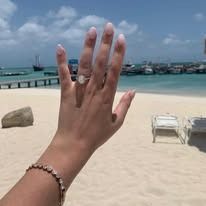 Hand showing off engagement ring on a beach with ocean and boats in the background.