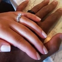 Hands intertwined; a white hand with an engagement ring and a black hand with a ring.