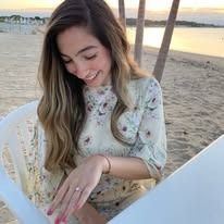 Woman with long hair smiles, wearing a floral dress, seated at the beach with a table.