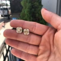 A hand holding two small, square, gold stud earrings with sparkling stones.
