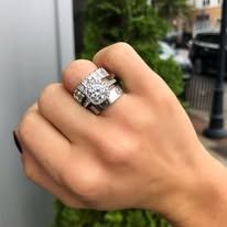 Person's hand with large diamond ring. Outdoor setting with green foliage, and blurred street in the background.