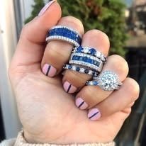 Hand wearing multiple rings with blue sapphires and diamonds; pink nail polish.