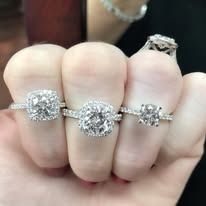 Close-up of three fingers each wearing a diamond engagement ring, plus one on the pinky finger.
