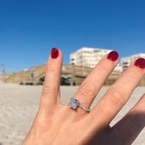Close-up of hand with a diamond ring on the beach, blue sky and buildings in the background.