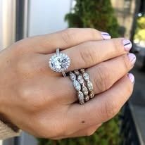 Hand displaying three diamond rings: engagement and two bands. Pink nails.