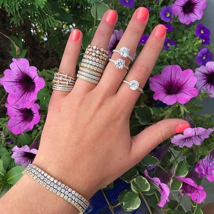 Hand with multiple diamond rings and bracelet, coral nails, purple flowers in background.