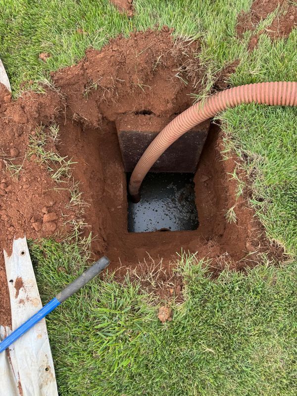 Open square hole in grass with a hose, presumably pumping water from a dark-colored concrete container.