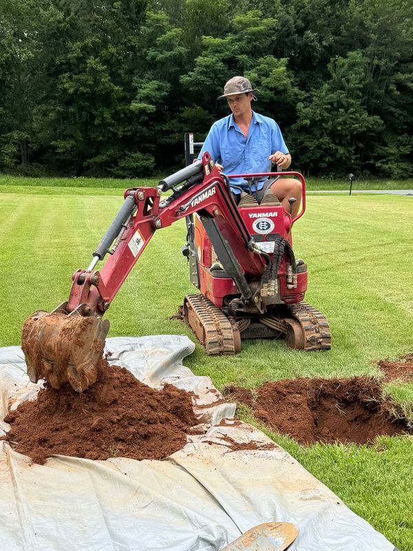 Man operating a small red excavator, digging in a grassy yard, with dark soil.