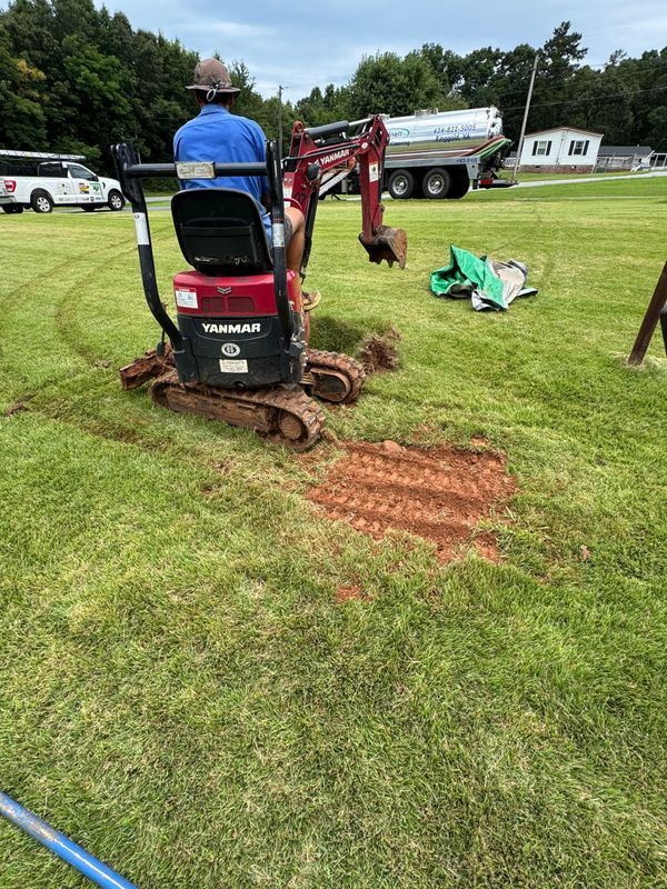 Person operating a Yanmar mini-excavator on a grassy field, digging a trench. A truck is parked nearby.