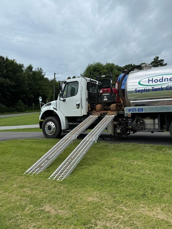 A white septic tank truck with ramps on grass. An excavator is being loaded.