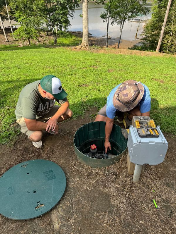 Two men working on a septic tank in a grassy yard near a lake. One kneels, the other leans in.