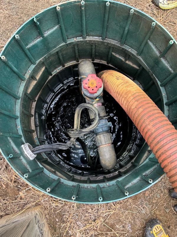 Open septic tank with dark liquid and orange hose. Green plastic casing; a valve and pipe visible.