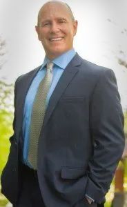 Bald man in blue suit, smiling with hand in pocket.