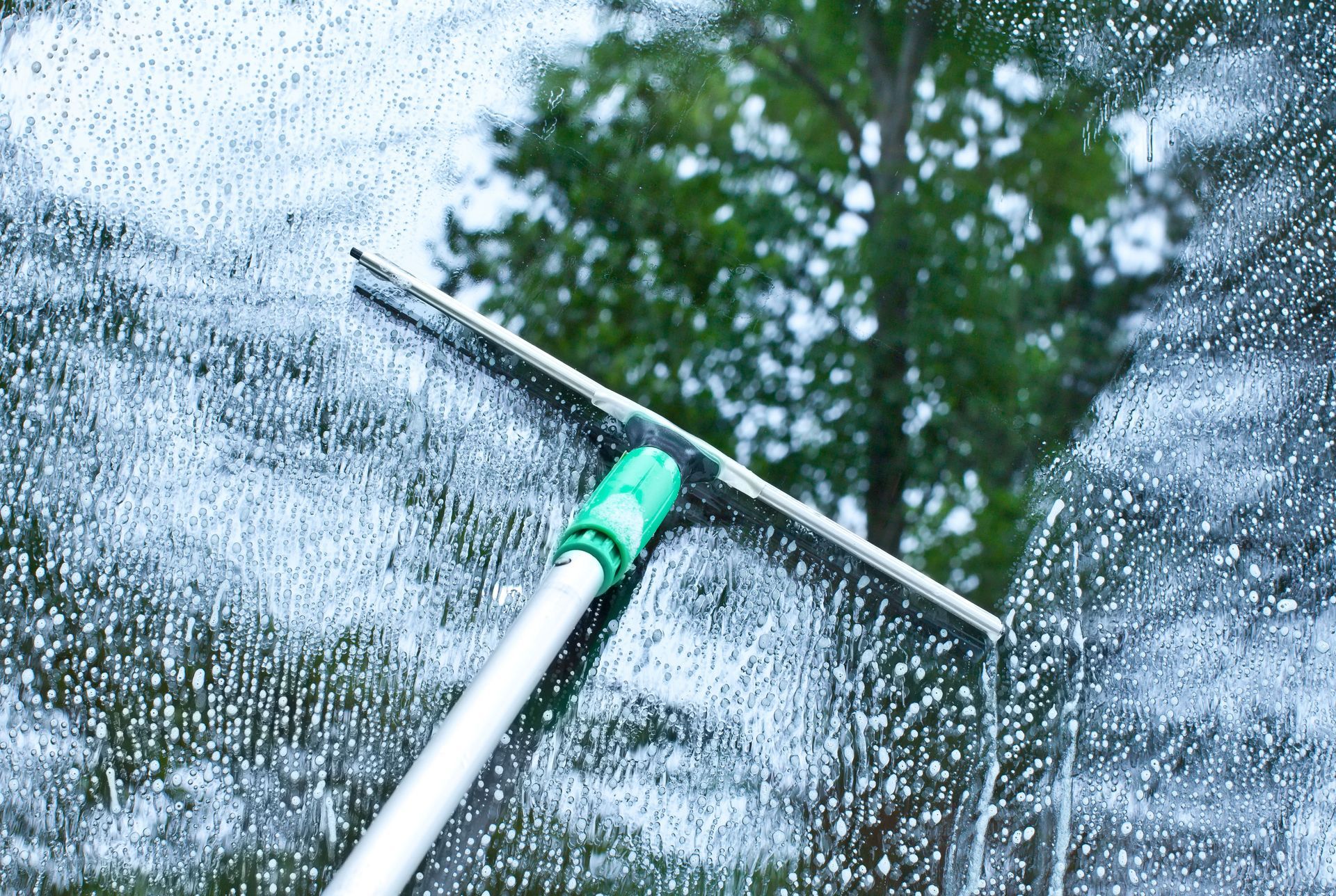 Window squeegee cleaning a wet glass pane outdoors amid water droplets and trees