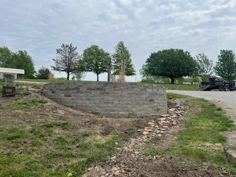 Stone retaining wall with shrubs, grass, and trees under a blue sky.