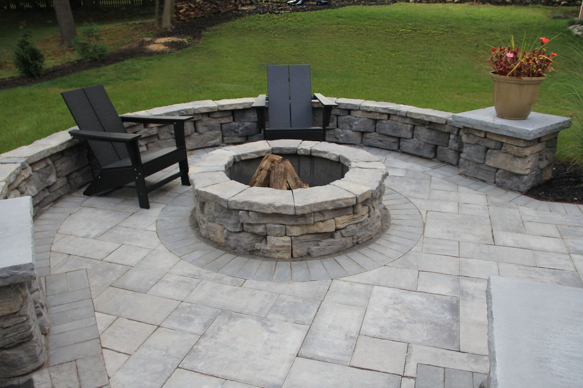 Stone patio with fire pit, seating, and a low stone wall in a grassy yard.