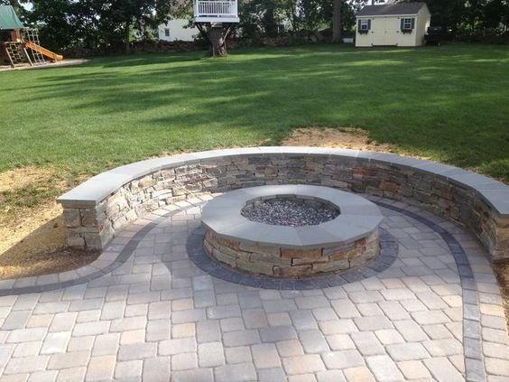 Stone fire pit surrounded by a stone wall and patterned brick patio in a grassy backyard.