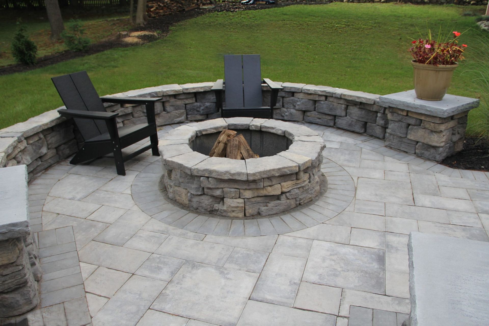 Stone patio with a fire pit, seating, and a low stone wall in a yard.