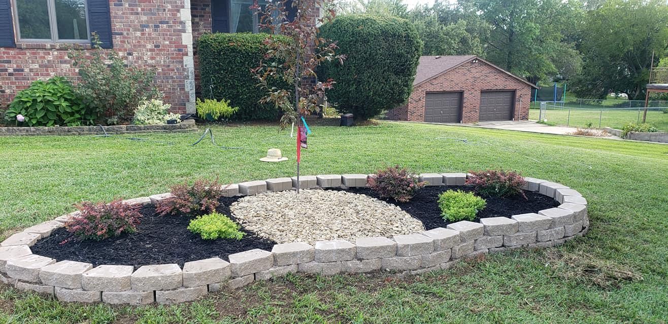 Oval-shaped landscaped garden bed in a yard, filled with mulch, stones, and plants, in front of a house.