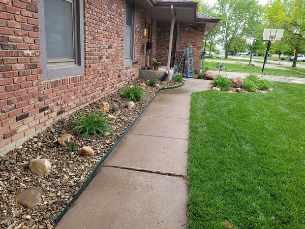 Brick house exterior with a concrete walkway, garden bed with plants, and green grass.