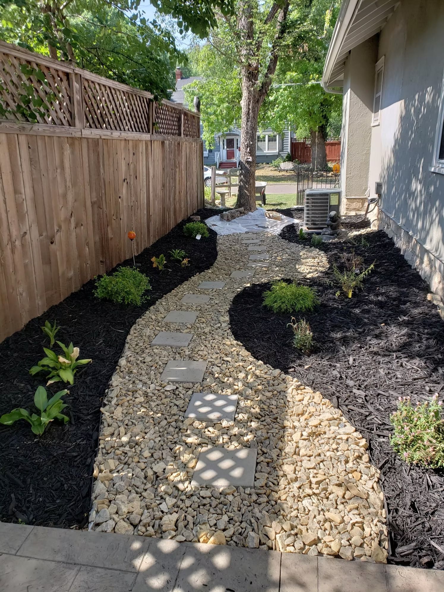 Stone pathway winds through a garden bed with mulch and plants alongside a wooden fence.