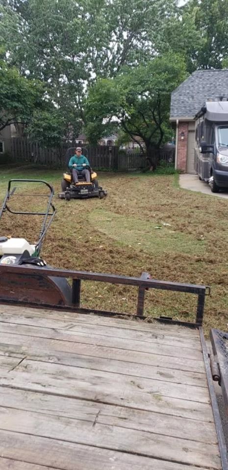 Person mowing lawn with riding mower, lawnmower and trailer in yard.