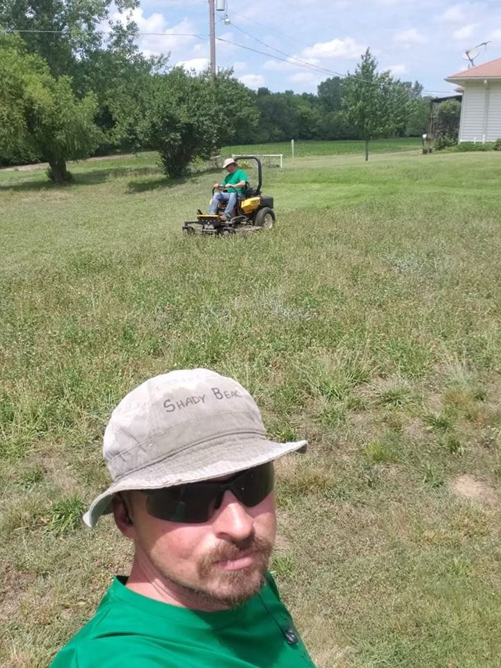 Man in hat takes selfie; another mows grass on riding mower in a sunny yard.