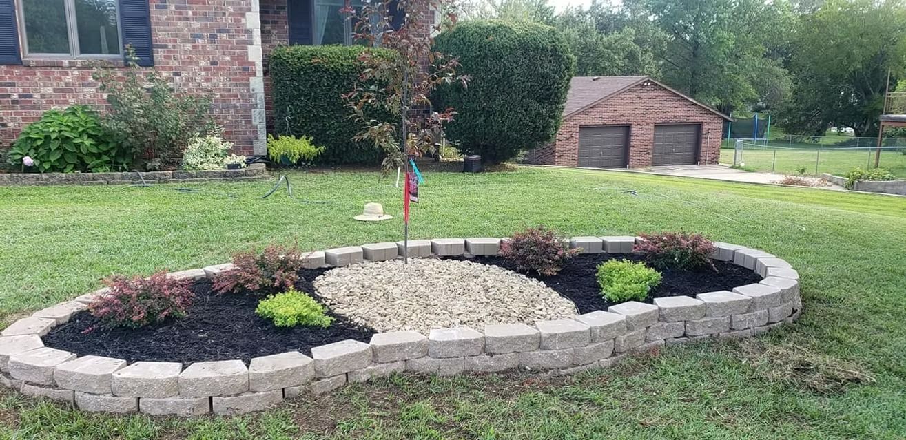 A landscaped front yard with a brick-lined flower bed filled with mulch, stones, and plants. A garage is in the background.