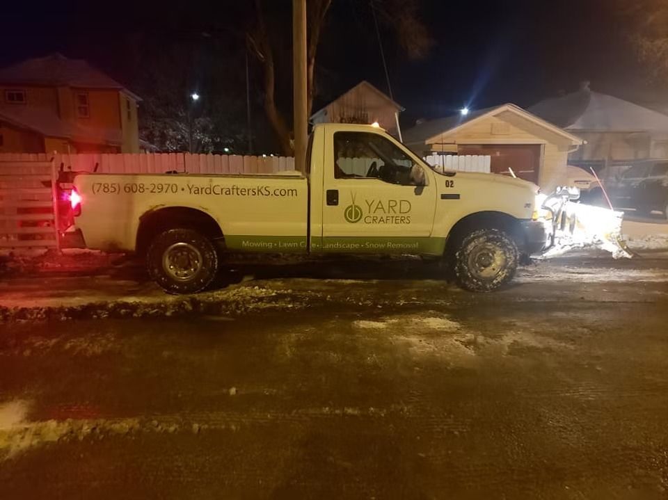 White pickup truck with snow plow plowing a snowy street at night.  