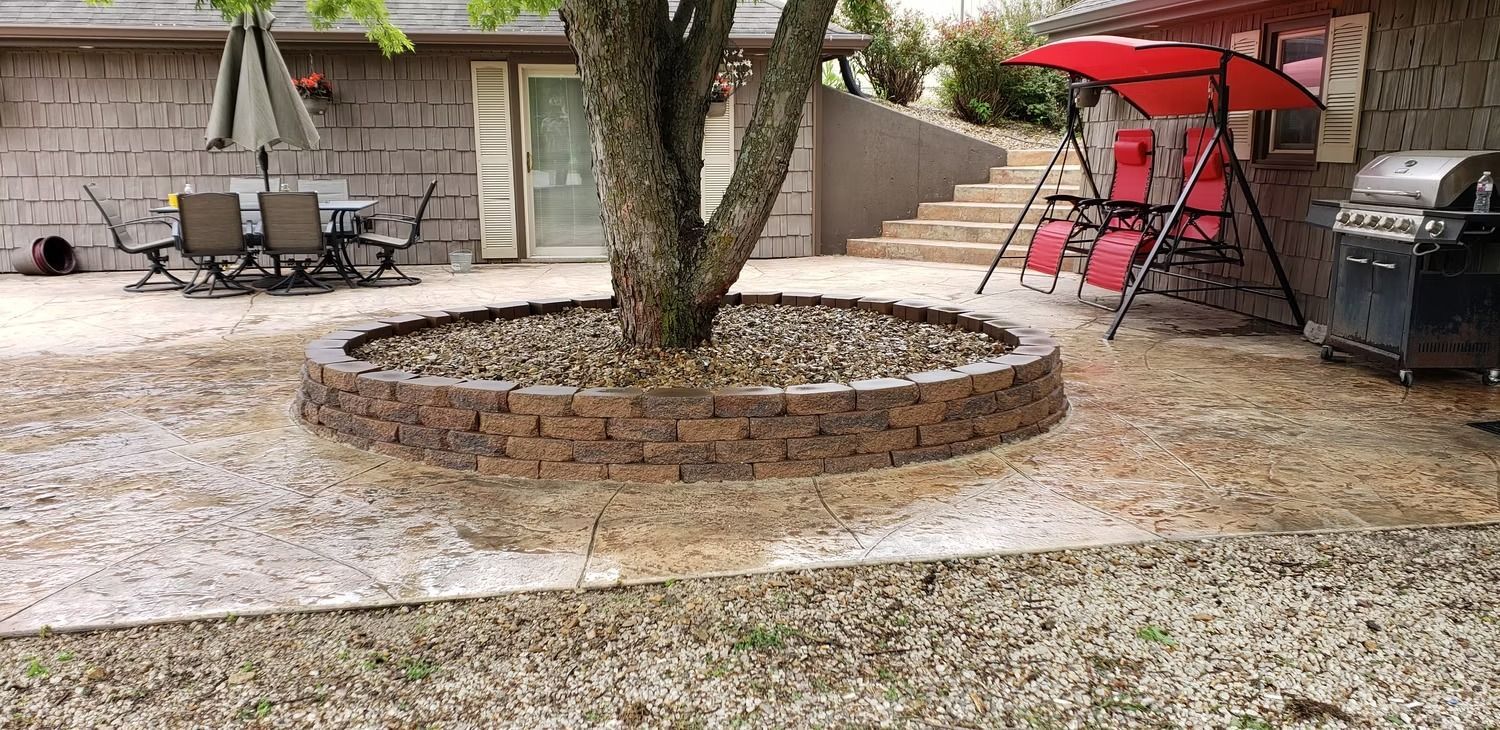 Backyard patio with a tree encircled by a stone border, gravel ground, and outdoor furniture.