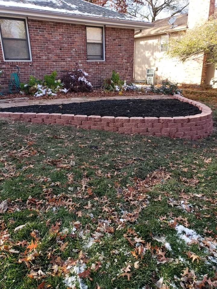 Brick-edged garden bed with dark mulch, partially snow-covered, in front of a brick house.