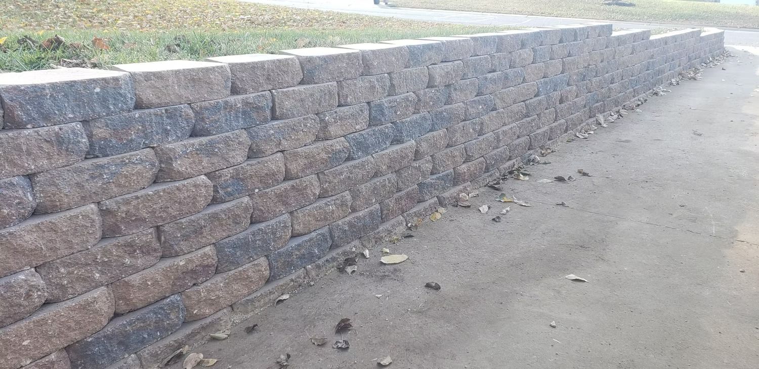 Stone retaining wall along a gray concrete surface. Green grass is visible in the background.