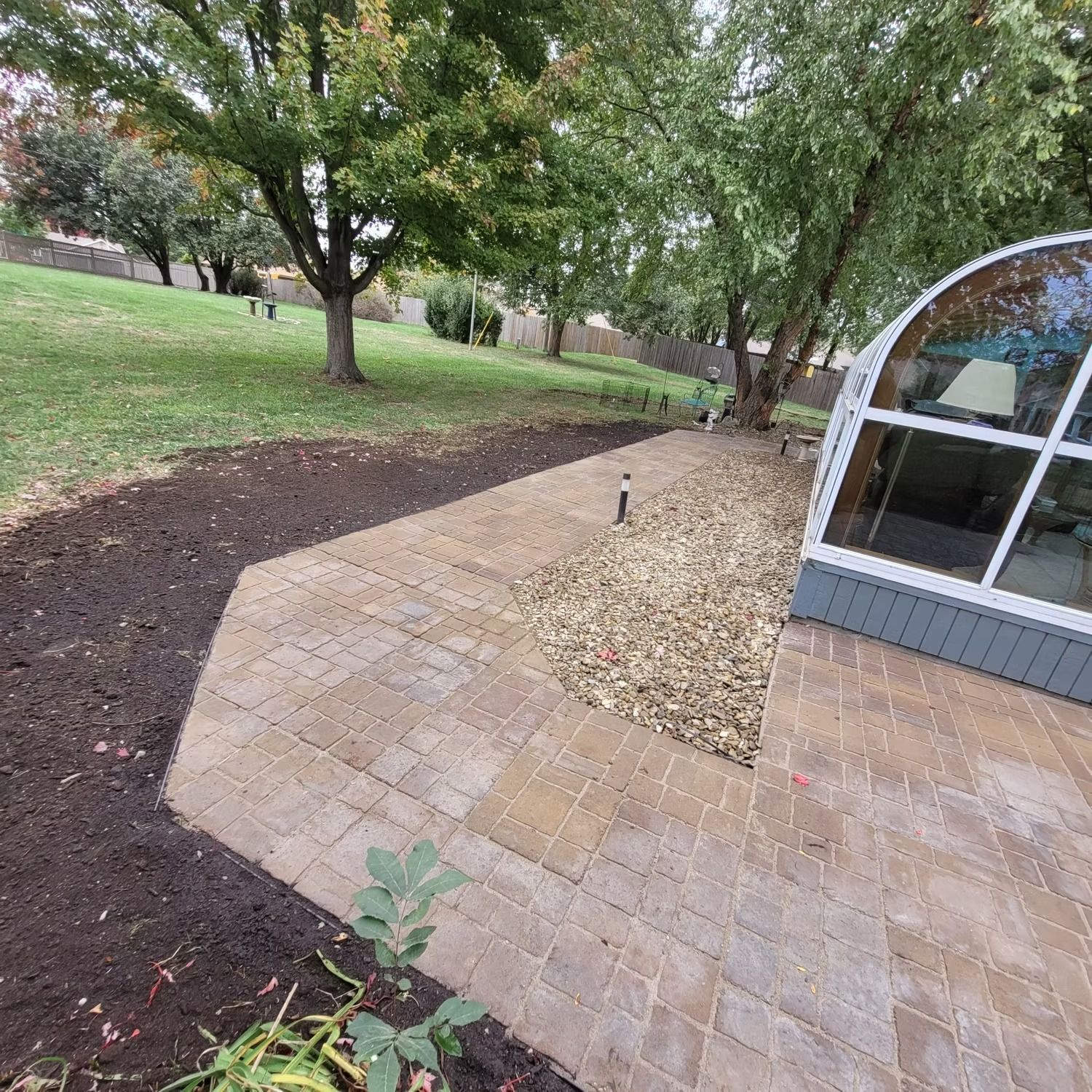 Stone patio with pathway, mulch, and trees. A building with a curved window is on the right.
