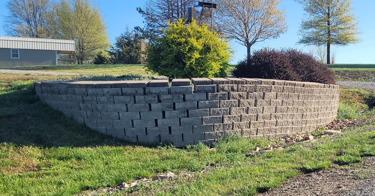 Stone retaining wall with shrubs, grass, and trees under a blue sky.