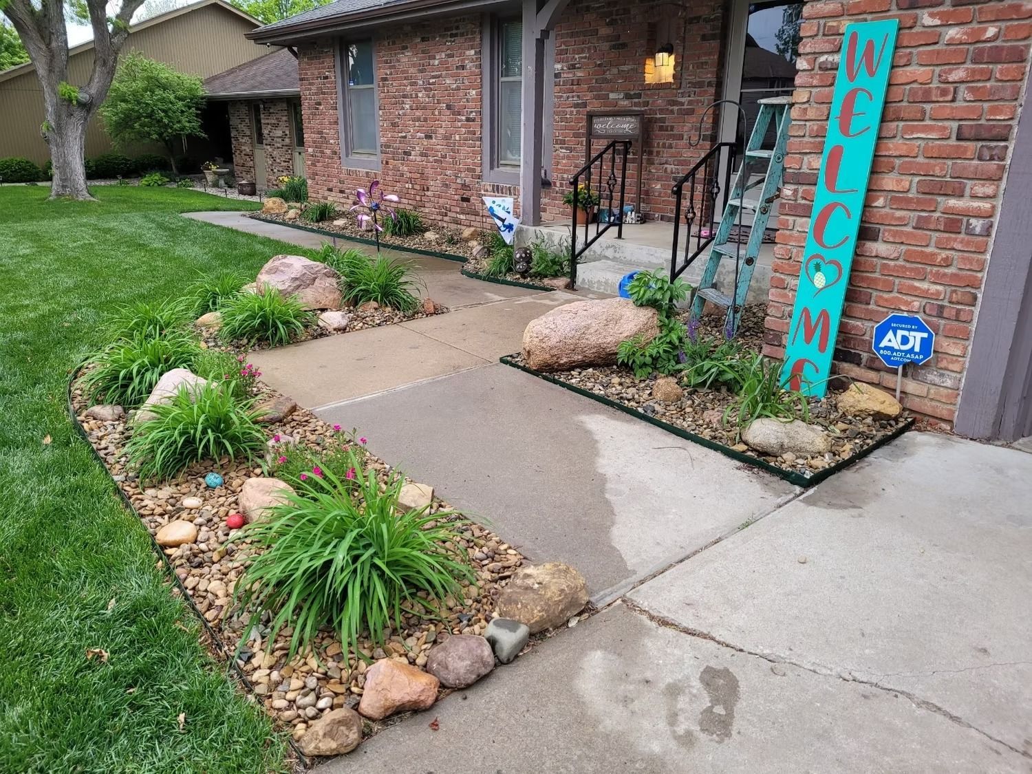 Brick house with a concrete walkway lined with rock gardens and greenery. A welcome sign stands by the entrance.