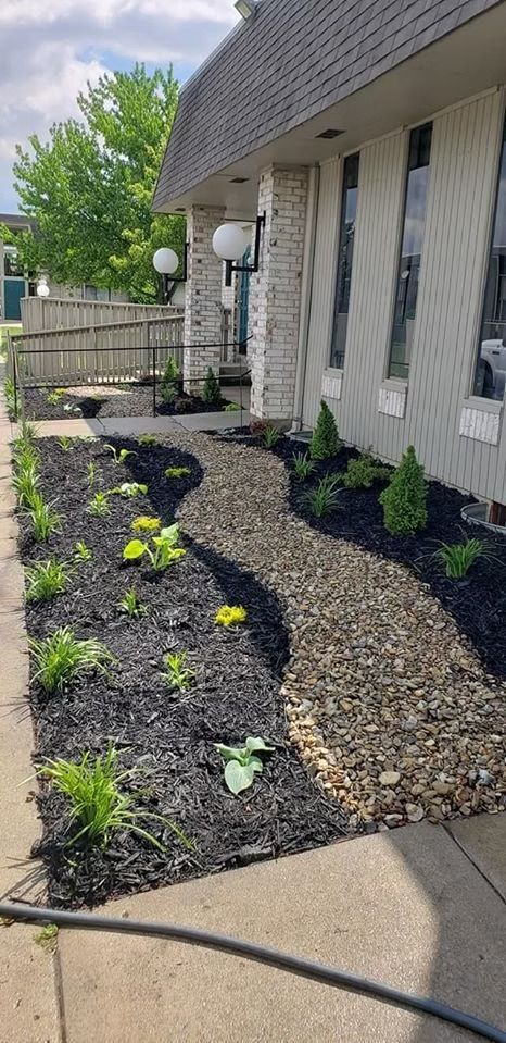 Pathway of pebbles winds through a landscaped bed, flanked by black mulch and plants, alongside a building.