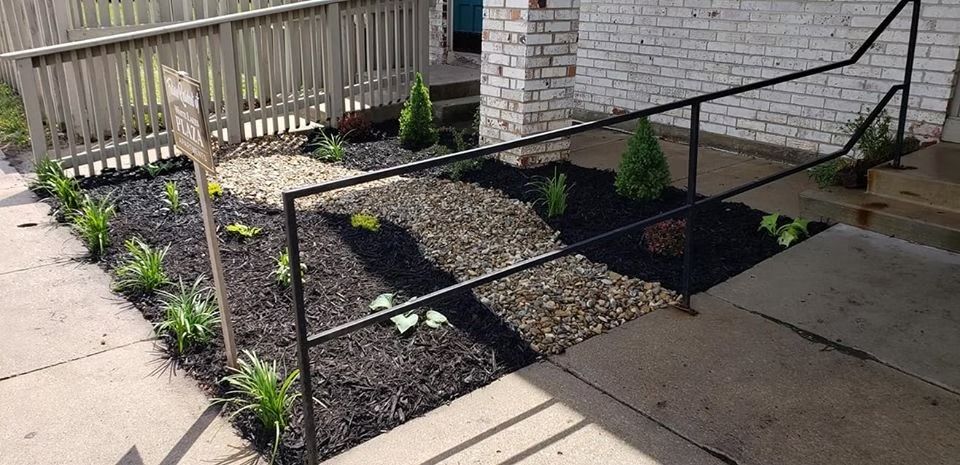 Small garden bed with black mulch, plants, and a black handrail next to a concrete walkway.